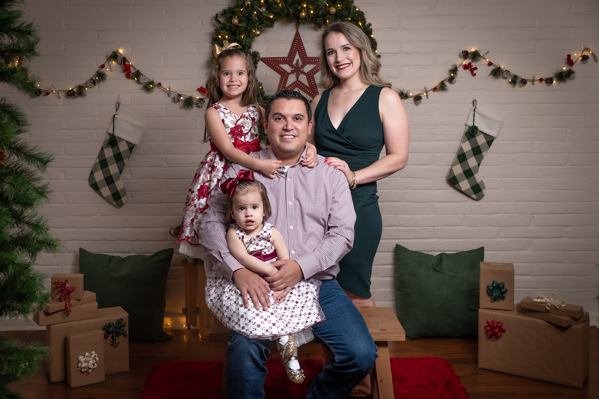 dad sitting on bench holding baby daughter whole mom and other daughter are standing and smiling at camera